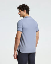 A curly-haired man models the Psycho Bunny MENS MONROE PIQUE POLO - B6K964D200 in light blue, featuring dark sleeve and collar trim. He stands gazing to the side against a plain white background, highlighting the polo's regular fit that balances comfort with style.