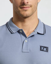 A man is seen from the shoulders up wearing a light blue Psycho Bunny MENS MONROE PIQUE POLO (B6K964D200) with dark collar trim, a logo appliqué patch on the chest, and a button-up collar in a regular fit.
