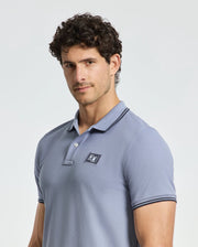 A man with curly hair is wearing a Psycho Bunny MENS MONROE PIQUE POLO - B6K964D200, in light blue with a small logo on the chest. He stands against a plain light gray background, looking slightly to the side.
