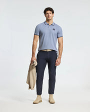 A man stands against a plain background wearing a light blue MENS MONROE PIQUE POLO by Psycho Bunny, paired with dark blue pants and beige shoes. Holding a light brown jacket, his neutral expression complements the polo's regular fit and subtle logo appliqué.