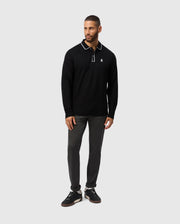 A man dressed in the Psycho Bunny MENS KENT LONG SLEEVE PIQUE POLO - B6M935D200, featuring a black color with white trim on the collar, stands against a gray background. His understated elegance is further enhanced by his dark pants and sleek black sneakers.