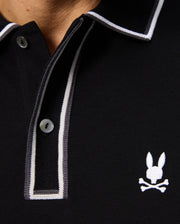 Close-up of the Psycho Bunny MENS KENT LONG SLEEVE PIQUE POLO - B6M935D200, a black rugby-style polo shirt made from soft Pima cotton. It features a white embroidered logo of a bunny with crossbones below it, exuding quiet luxury. The design is enhanced by its two-button placket and contrasting white and gray trim along the collar.