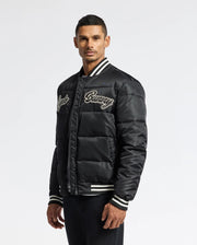 A man wearing a Psycho Bunny MENS FRASER FASHION PUFFER - B6N338E200 in black, featuring white striped cuffs and an embroidered script on the chest. The background is plain white.