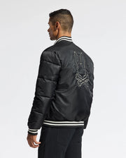 A person stands facing away, wearing the MENS FRASER FASHION PUFFER by Psycho Bunny. This stylish black bomber jacket features a rabbit and crossed bones design on the back, with white striped cuffs and collar, providing both style and comfort against the plain white background.
