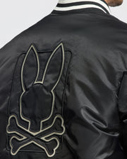 A person is wearing the stylish black MENS FRASER FASHION PUFFER by Psycho Bunny. This warm jacket features a striking large embroidered design on the back, depicting a stylized skull and crossbones with bunny ears. It also boasts a black and white striped collar, effortlessly blending warmth with edgy fashion.