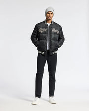 A man wearing a gray beanie and a black bomber jacket with "Richie" and "Savage" text stands in the studio. He showcases a stylish look with the MENS FRASER FASHION PUFFER by Psycho Bunny, paired with black pants and white sneakers, his hands casually tucked into his pockets against a plain backdrop.