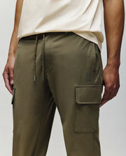 A person wearing Psycho Bunny MENS LLOYD CARGO JOGGER - B6P159B200 in olive green, featuring side pockets, drawstring waist, and detachable keychain, paired with a light beige t-shirt. Only the lower torso and upper legs are visible.