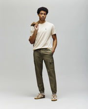 A man stands against a plain background wearing Psycho Bunny's MENS LLOYD CARGO JOGGER - B6P159B200, a beige t-shirt, tan sneakers, and holds a brown jacket over his shoulder, gazing straight ahead with a neutral expression.