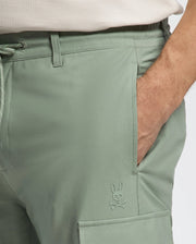 A person wearing Psycho Bunny's Mens Lloyd Cargo Jogger - B6P159B200 in light green stretch twill, featuring an embossed bunny and crossbones logo above the front flap pocket, with one hand in the pocket. They also wear a light-colored shirt.