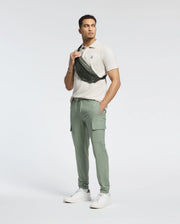 A man in a beige polo, Psycho Bunny's MENS LLOYD CARGO JOGGER - B6P159B200, and white sneakers stands against a white background. He carries an embroidered stylish black crossbody bag.