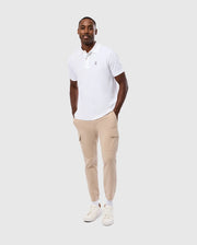 A person stands smiling with hands in their snap fastening pockets, wearing a white polo shirt, Psycho Bunny MENS LLOYD CARGO JOGGER (B6P159B200) in beige, and white sneakers. The background is plain and light gray.