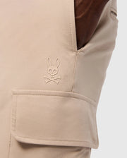 Close-up of a pocket on the MENS LLOYD CARGO JOGGER - B6P159B200 by Psycho Bunny, showcasing a stitched logo of a bunny head with crossbones underneath. The smooth fabric pocket has a flap secured by snap fastening, and a hand is casually resting in the side pocket, highlighting the refined embroidery.