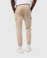 A person is shown from the waist down, wearing a white shirt, white sneakers, and the MENS LLOYD CARGO JOGGER - B6P159B200 by Psycho Bunny. The beige joggers feature snap fastening pockets and have elastic cuffs at the ankles. The person’s hands are placed in their pockets against a plain white background.