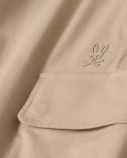Close-up of the MENS LLOYD CARGO JOGGER - B6P159B200 by Psycho Bunny, featuring a beige fabric adorned with a snap fastening pocket. Above the pocket is a stitched bunny logo with crossed bones underneath its head, showcasing a subtle pirate motif. The playful embroidery refinement adds an elegant touch to the smooth and well-crafted fabric.