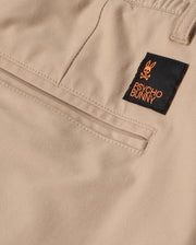 Close-up of the beige fabric MENS LLOYD CARGO JOGGER - B6P159B200 by Psycho Bunny, showcasing an orange-accented black label with a bunny logo and the words "Psycho Bunny" stitched on. The jogger pants exhibit detailed stitching and refined embroidery around the snap fastening pockets.