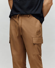 A person wears Psycho Bunny MENS LLOYD CARGO JOGGER - B6P159B200, tan cargo joggers with drawstring waist, side pockets, and subtle embroidery, paired with a black shirt and hands tucked behind their back.