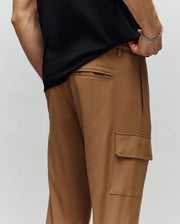 A person wears Psycho Bunny MENS LLOYD CARGO JOGGER - B6P159B200 in light brown, shown from the waist down with one hand in their pocket. The joggers have a back pocket, side cargo pocket, and subtle embroidery detail.