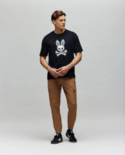 A man stands against a plain background wearing a black Psycho Bunny T-shirt with a white bunny and crossbones, MENS LLOYD CARGO JOGGER - B6P159B200 pants, and black athletic sneakers with white stripes.
