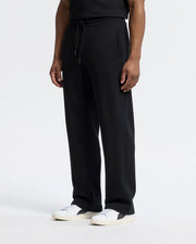 The person is wearing Psycho Bunny MENS LYNX WIDE LEG CLASSIC FRENCH TERRY SWEATPANT - B6P379E200 in black, paired with white sneakers. Their arms rest naturally by their sides against a plain white background.