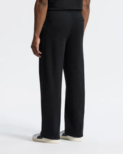 The image features the back view of a person wearing Psycho Bunny's MENS LYNX WIDE LEG CLASSIC FRENCH TERRY SWEATPANT in black and black and white sneakers. An embroidered logo subtly enhances their style against a plain white background.