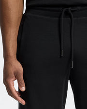 A person wears Psycho Bunny's MENS LYNX WIDE LEG CLASSIC FRENCH TERRY SWEATPANT - B6P379E200 in black, with their left hand near the pocket. The light neutral background highlights the fabric’s soft texture and the drawstring's metal tips.