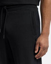 A close-up shows a person in Psycho Bunny's Mens Lynx Wide Leg Classic French Terry Sweatpants (B6P379E200), featuring a drawstring and embroidered logo near the pocket. The individual rests their right hand on their thigh while wearing a matching black shirt.