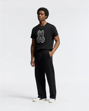 A man wears a Psycho Bunny black T-shirt with a bunny and crossed bones graphic, paired with the MENS LYNX WIDE LEG CLASSIC FRENCH TERRY SWEATPANT and white shoes, standing against a plain background and looking off to the side with a neutral expression.