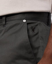 A close-up shows someone in Psycho Bunny's MENS GILMAN CHINO PANT - B6P393Z1WB, dark green with an embroidered bunny logo near the front pocket. The comfort suits workdays to weekends, paired with a tucked-in light shirt. A logo metal button is visible on the waistband.