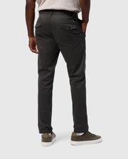 A person in Psycho Bunny MENS GILMAN CHINO PANT - B6P393Z1WB is seen from behind against a light gray backdrop, wearing dark gray chinos with back pockets and olive green sneakers, hands tucked in pockets, effortlessly transitioning from workday to weekend style.