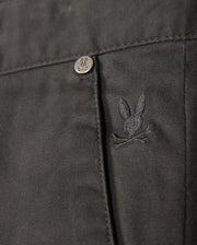 The Psycho Bunny MENS GILMAN CHINO PANT - B6P393Z1WB features a close-up of fabric with an embroidered rabbit head and crossed bones logo. A metal button displays the same design. This dark, comfy chino transitions smoothly from workday to weekend, making it a versatile wardrobe staple.