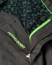 Close-up of the MENS GILMAN CHINO PANT - B6P393Z1WB by Psycho Bunny in dark gray, featuring "Psycho Bunny" embroidered in neon green on the pocket. Designed for comfort from workday to weekend, with an inner lining of outlined bunnies and a partially open zipper showing more green accents.