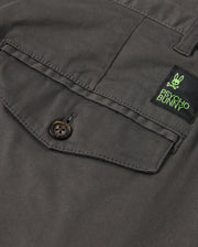 Close-up of a dark gray shirt pocket with button fastening. Above it, a black and green label features a bunny design and "Psycho Bunny." Pairing perfectly with the MENS GILMAN CHINO PANT - B6P393Z1WB from Psycho Bunny, this classic style smoothly transitions from workday to weekend.