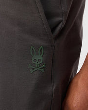 Close-up of a person wearing dark-colored Psycho Bunny MENS MADISON COMMUTER PANT - B6P395Z1WB. The pants feature a small green skull and crossbones logo with bunny ears near the pocket, showcasing subtle Bunny detailing. The person's arm is partially visible on the right side of the image.