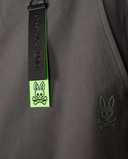 Close-up of a dark MENS MADISON COMMUTER PANT - B6P395Z1WB featuring a black and neon green zipper pull with the Psycho Bunny logo—a bunny skull and crossbones. The logo, embroidered in green near the zipper pull, complements the detachable Bunny keychain for added flair.
