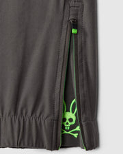 The image displays a close-up view of a dark-colored garment with a partially opened vertical zipper, revealing the bright green fabric of the MENS MADISON COMMUTER PANT - B6P395Z1WB by Psycho Bunny. The green fabric showcases a graphic design featuring a skull with rabbit ears and crossbones, incorporating distinctive bunny detailing.