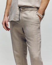 A person wears Psycho Bunny's MENS FATE LINEN DRAWSTRING PANT (B6P471C200) in light beige with a matching shirt, styled simply with a beaded bracelet and embroidered logo, standing against a plain neutral background.