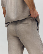 A person models the Psycho Bunny MENS FATE LINEN DRAWSTRING PANT (B6P471C200) in light beige, with a matching shirt. One hand rests in their pocket, highlighting the back pocket and side. The plain, light background keeps focus on the pants.