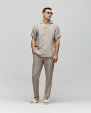 A man wearing Psycho Bunny's MENS FATE LINEN DRAWSTRING PANT (B6P471C200) in beige, a matching linen blend short-sleeve shirt, and beige slide sandals stands against a plain light background with his hands in his pockets.
