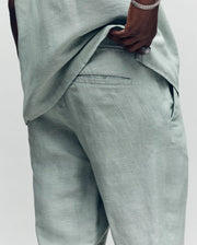 A man in light sage green Psycho Bunny MENS FATE LINEN DRAWSTRING PANT B6P471C200 lifts his matching shirt to show the back pocket with an embroidered logo. He's accessorized with a silver bracelet and ring.