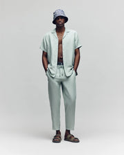 A man stands against a plain background, wearing Psycho Bunny’s MENS FATE LINEN DRAWSTRING PANT (B6P471C200) with a light gray linen-blend short-sleeve button-up shirt, a patterned bucket hat, sandals, and his hands in his pockets.