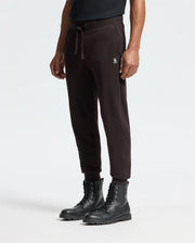 A person is wearing the Psycho Bunny Men's Levy Essential Fleece Sweatpant (B6P721D200) in black, made of 100% cotton with a small embroidered logo on the left side, paired with black lace-up combat boots. This casualwear look stands out against the plain white background.