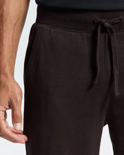 A person is wearing Psycho Bunny's MENS LEVY ESSENTIAL FLEECE SWEATPANT in black, with one hand resting at the side. This casualwear boasts pockets and a ribbed waistband for ultimate comfort, set against a plain and light-colored background.