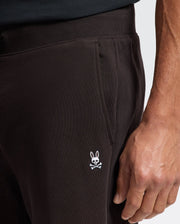 A person dressed in the MENS LEVY ESSENTIAL FLEECE SWEATPANT - B6P721D200 by Psycho Bunny, featuring a subtle embroidered bunny skull and crossbones emblem on the side. Their hand casually rests near the pocket, perfectly matching their dark top.