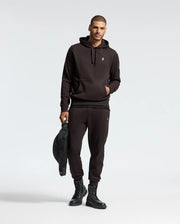 A man wearing a black hoodie and the MENS LEVY ESSENTIAL FLEECE SWEATPANT by Psycho Bunny stands against a plain background, holding a black bag. He is also sporting black lace-up boots and has one hand in his pocket, capturing the essence of casualwear.