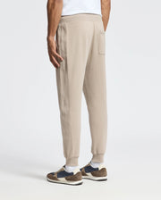 A person wearing the MENS LEVY ESSENTIAL FLEECE SWEATPANT in beige by Psycho Bunny and a white shirt, viewed from the back. They complete their look with dark blue and brown sneakers against a plain white background.