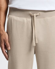 A close-up image features a person wearing MENS LEVY ESSENTIAL FLEECE SWEATPANT - B6P721D200 by Psycho Bunny, crafted from ultra-soft cotton fleece with a drawstring, paired with a white T-shirt. The person's left hand rests near their pocket, exuding comfort against the plain, light background.
