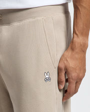 A close-up of someone wearing Psycho Bunny's MENS LEVY ESSENTIAL FLEECE SWEATPANT - B6P721D200, which are made from ultra-soft cotton fleece. The beige joggers display a small embroidered rabbit with crossed bones logo and have their hand nestled in the pocket. The look is finished off with a crisp white shirt.