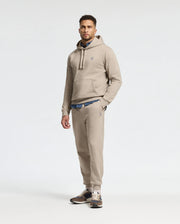 A man stands against a plain white background wearing the MENS LEVY ESSENTIAL FLEECE SWEATPANT in beige, crafted from ultra-soft cotton fleece by Psycho Bunny. Beneath his hoodie, a blue shirt adds a pop of color to the ensemble, while sneakers complete this effortlessly stylish look.