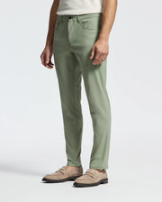 A person wearing Psycho Bunny's MENS MAVERICK 10K STEP PANT - B6P788D200 in light green, with five-pocket styling and slim-fit design, stands hands at their sides. They pair the pants with a white shirt and light brown loafers against a plain white background.
