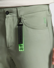 A close-up shows a person wearing light green Psycho Bunny MENS MAVERICK 10K STEP PANT with five-pocket styling and a dark green strap on the belt loop. The strap features a bunny logo and "Psycho Bunny" text. The pants are crafted from four-way stretch fabric, with a small bunny logo near the pocket.